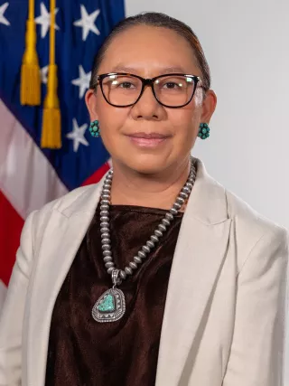 Headshot of woman with brown hair and glasses standing in front of an American flag.