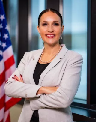 A woman with dark hair in a low bun dressed in professional clothing smiles.