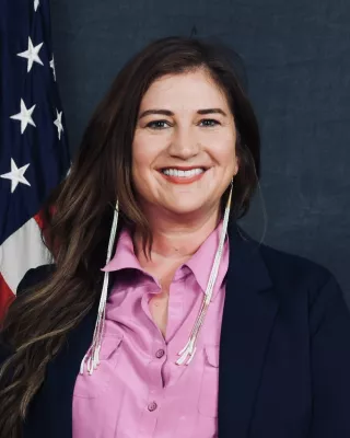 A woman with long brown hair in a button up shirt and blazer stands in front of an American flag for a headshot.