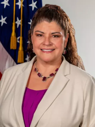 A headshot of a woman with long brown hair in a pony tail wearing a beige blazer and pink top.