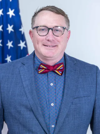 A man in a blue suitcoat and red bowtie poses for a photo in front of an American flag.