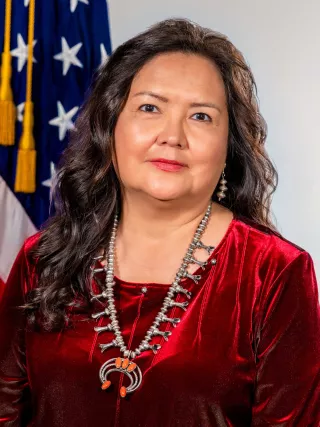 A headshot of a woman with long, curly brown hair in a red top in front of an American flag.