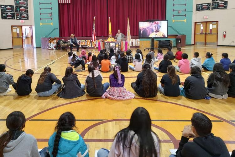 Children sit in lines on a gym floor and listen to a veteran discuss his service.