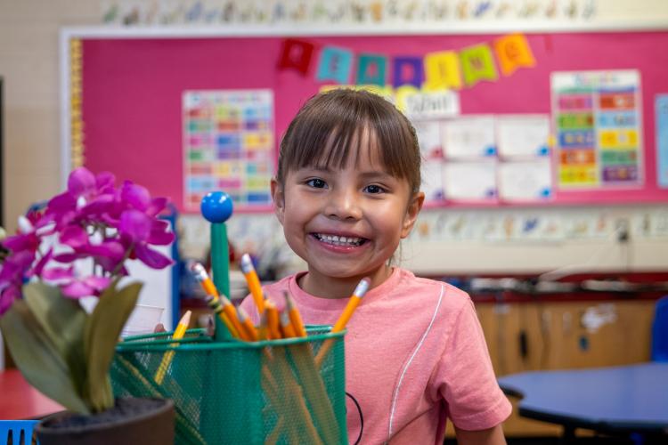 A student stands in a classroom with a container of pencils and flowers in front of her.