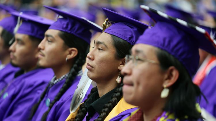 College students in purple graduation caps and gowns sit at a graduation ceremony and face forward.