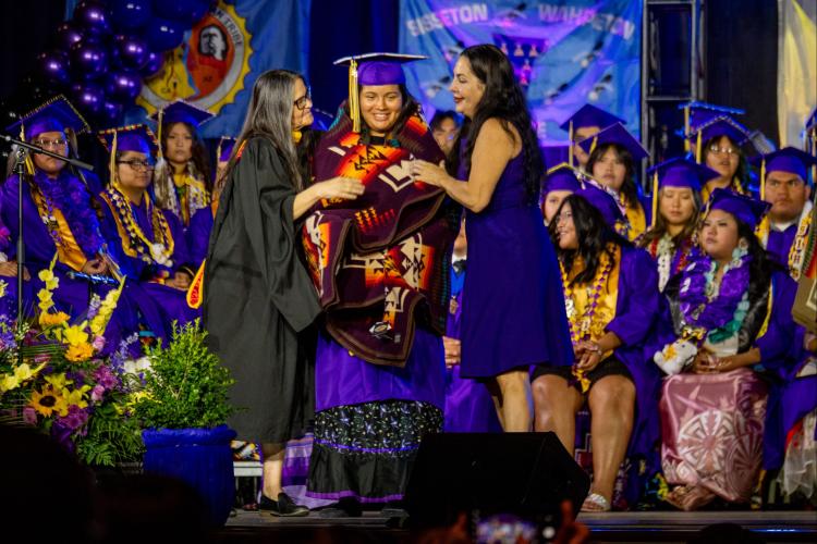A student in a purple graduation robe and cap stands center stage between two teachers who wrap a blanket around her.