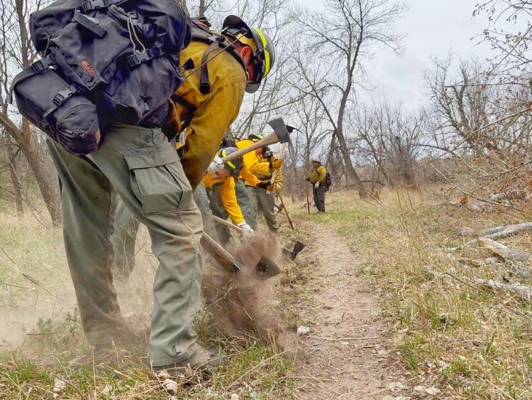 Students in firefighting gear practice hitting into the soil with an axe outdoors.