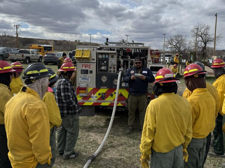 A group of students in firefighter gear stand looking toward a fire truck and listen to someone present.