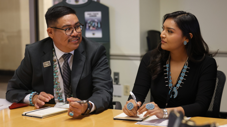 Two students - one male, one female - sit at a table while dressed professionally and listen to each other talk and write in notebooks.