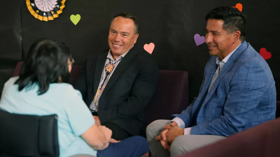 Three people sit on couches and talk while smiling.