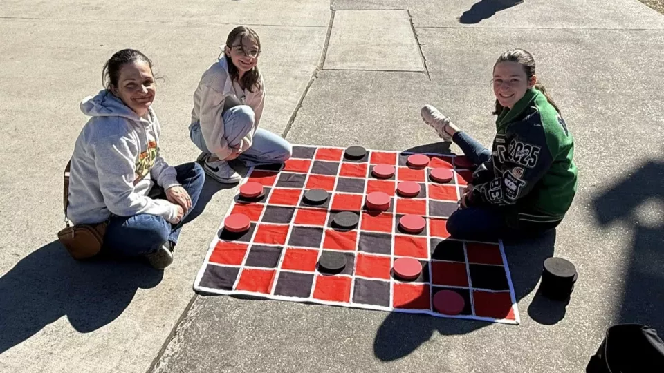 Three children sit on the ground and play giant checkers.