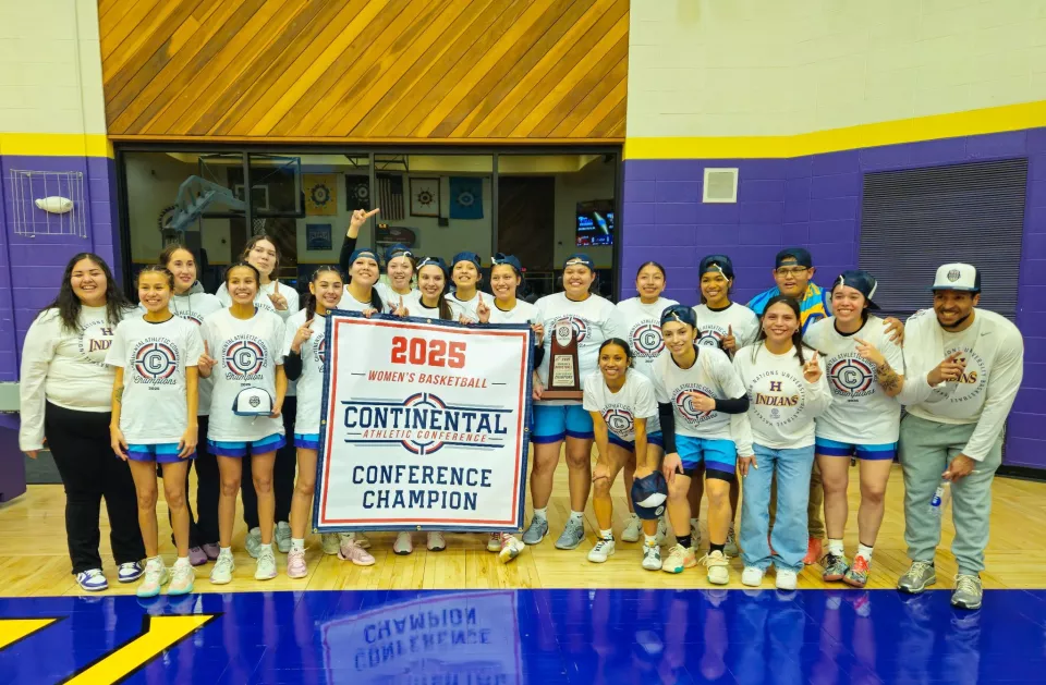 A women's basketball team poses together in a gymnasium, celebrating their championship victory. They wear matching white shirts with "Continental Athletic Conference Champions 2025" printed on them. The team members hold a large banner that reads "2025 Women's Basketball Continental Athletic Conference Champion," along with a trophy. Many of the players are smiling, some pointing upwards, and a few wearing their caps backward. Coaches and supporters stand alongside them. The gymnasium has purple and yellow