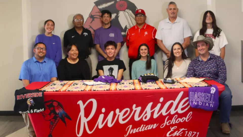 A group of 12 people sit and stand behind a table as two students in the middle sign papers.