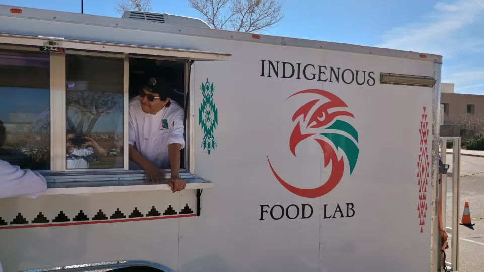A man in a white chef's coat stands in the window of a food truck that reads "Indigenous Food Lab."