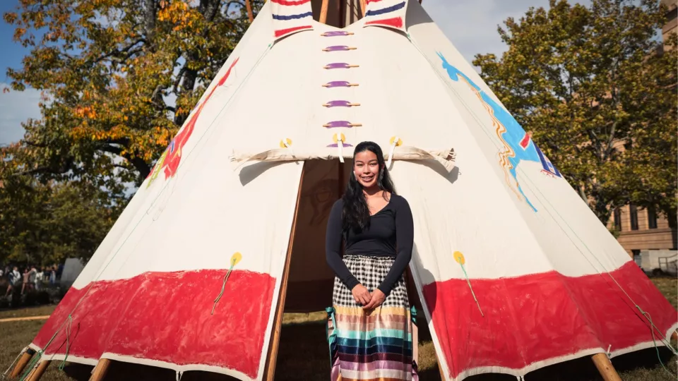 A woman in a ribbon skirt stands at the entrance of a painted tipi with trees in the background.