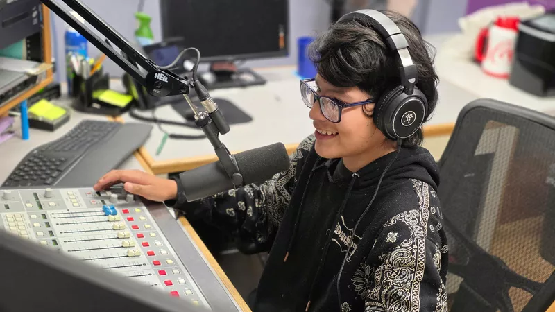 A student wearing glasses and headphones sits in front of a microphone at an audio mixing board.