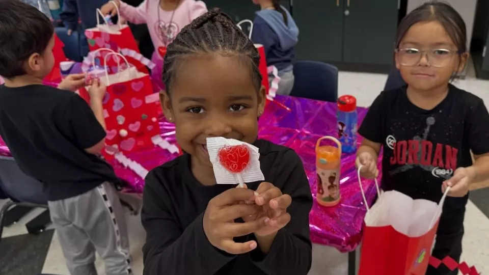 A young student smiles while holding up a red, heart-shaped lollipop.