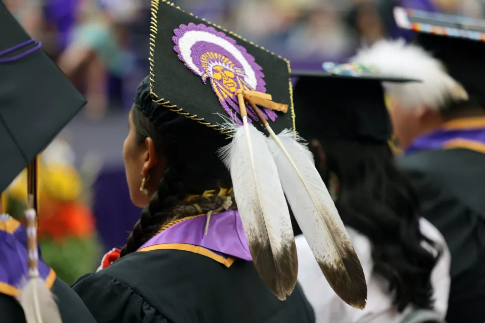 A close-up view of Native American graduates in caps and gowns at a graduation ceremony. The central graduate wears a decorated cap featuring a detailed beaded design of a Native figure in purple, white, and gold, bordered with gold thread and adorned with two large white feathers hanging down the back. Other graduates nearby also wear traditional regalia elements with their academic attire. The scene conveys pride, cultural identity, and academic achievement.