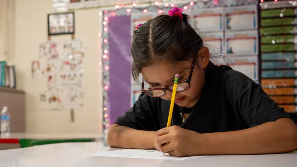 A student sits at her desk and writes on a worksheet with a pencil.