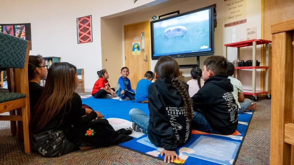 Students sit in a circle on a classroom floor and look up to watch a video on a TV.