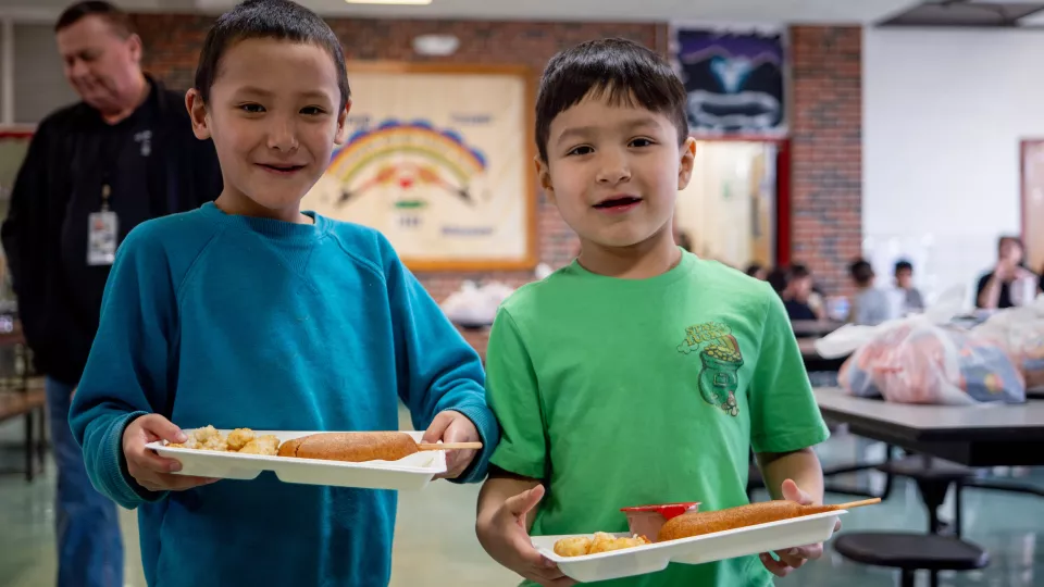 Two young students stand in a cafeteria holding their lunch trays with food.