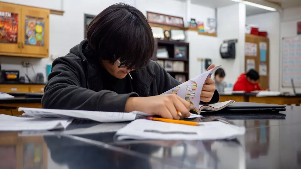 A student wearing glasses sits at a lab table in a classroom and looks at a science textbook and worksheets.