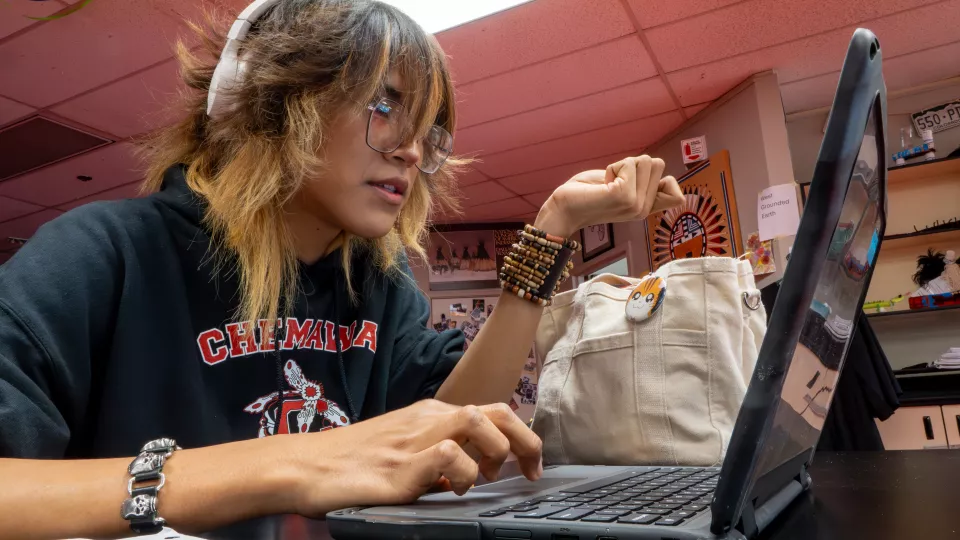 A student wearing headphones sits at a desk looking at their laptop.