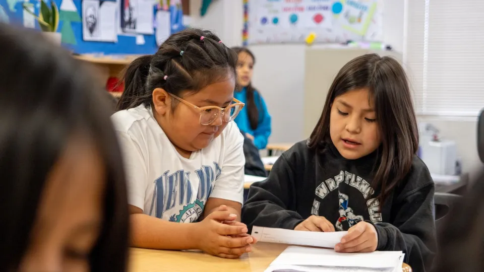 Two female students sit at a desk together and look at a piece of notebook paper.