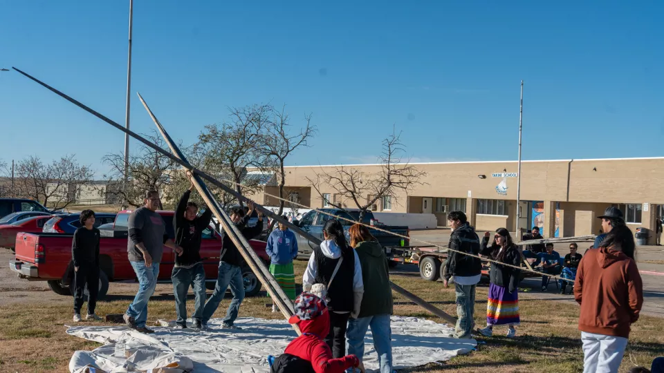 A group of students and adults work together to raise up a tipi on a lawn.