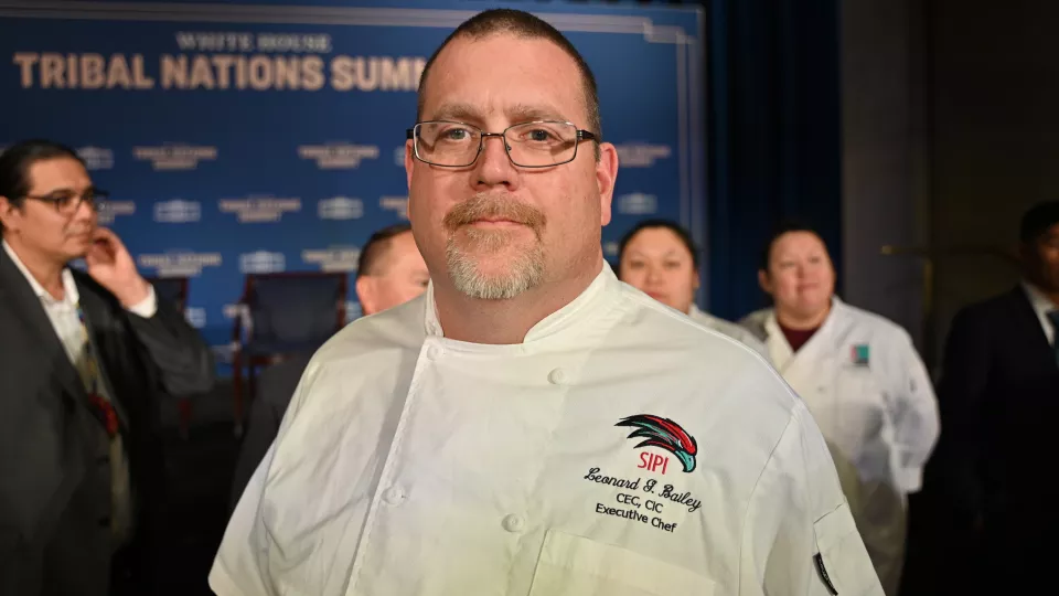 A man in a chef's coat with a goatee, buzzed hair and glasses.