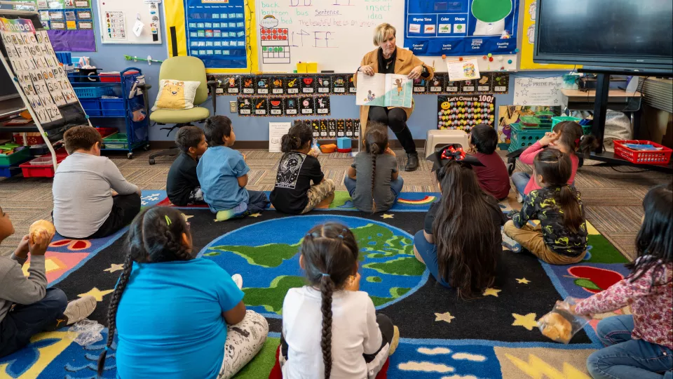 A group of students listen to their teacher read them a book while sitting on the floor.