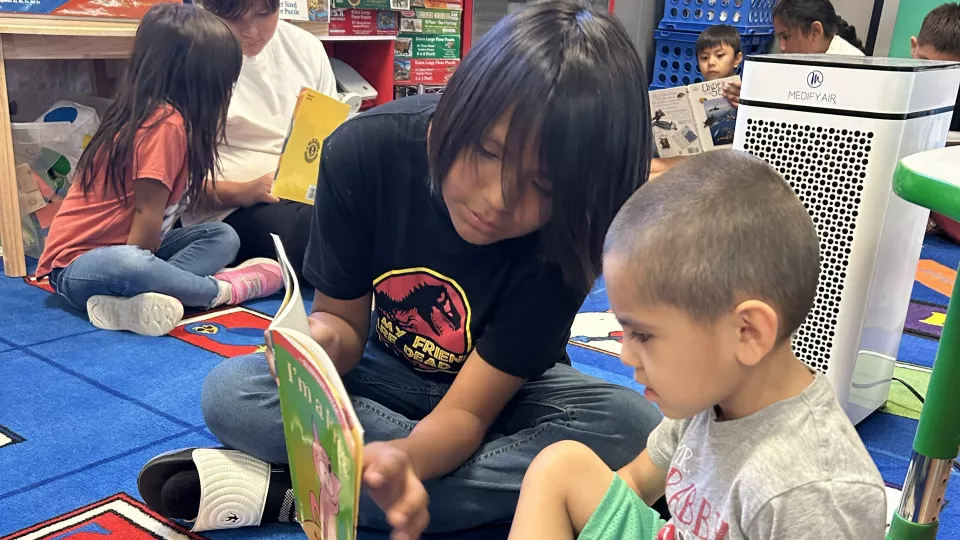 An older elementary student sits on a rug and reads with a first grader.