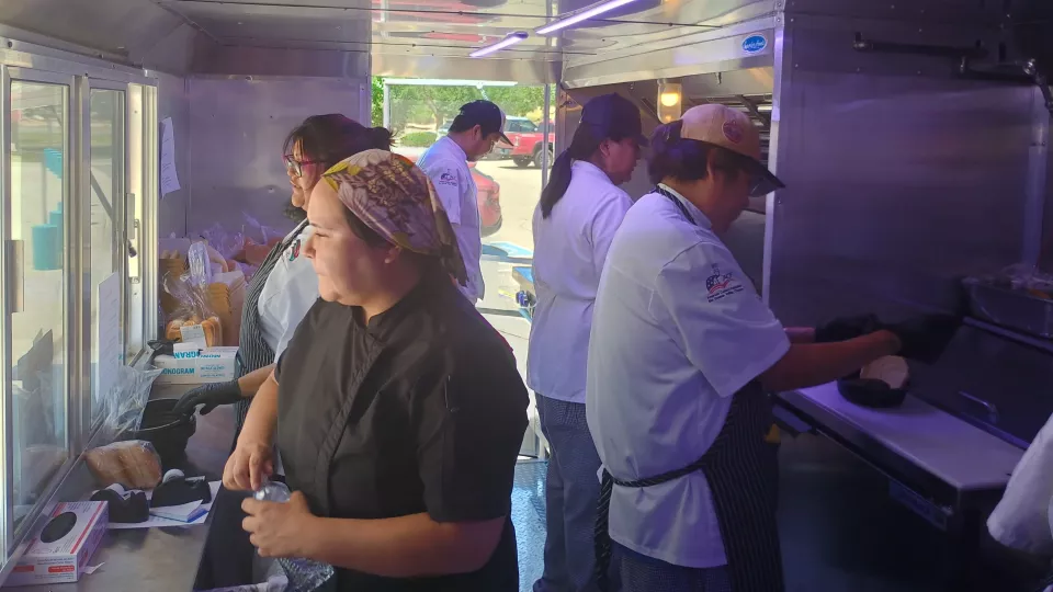 Five students in chef coats stand inside a food truck and prepare and serve food.