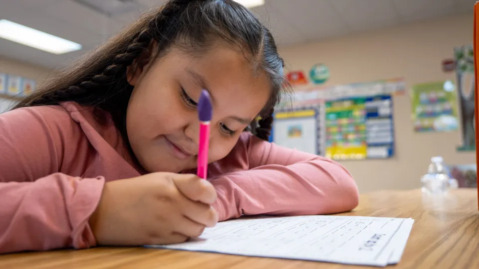 A young female student with braids sits at a desk and writes with a hot pink pencil.