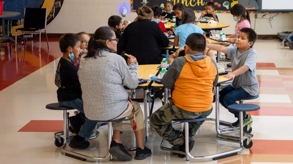 Students and staff sit and enjoy lunch at a round table in a cafeteria.