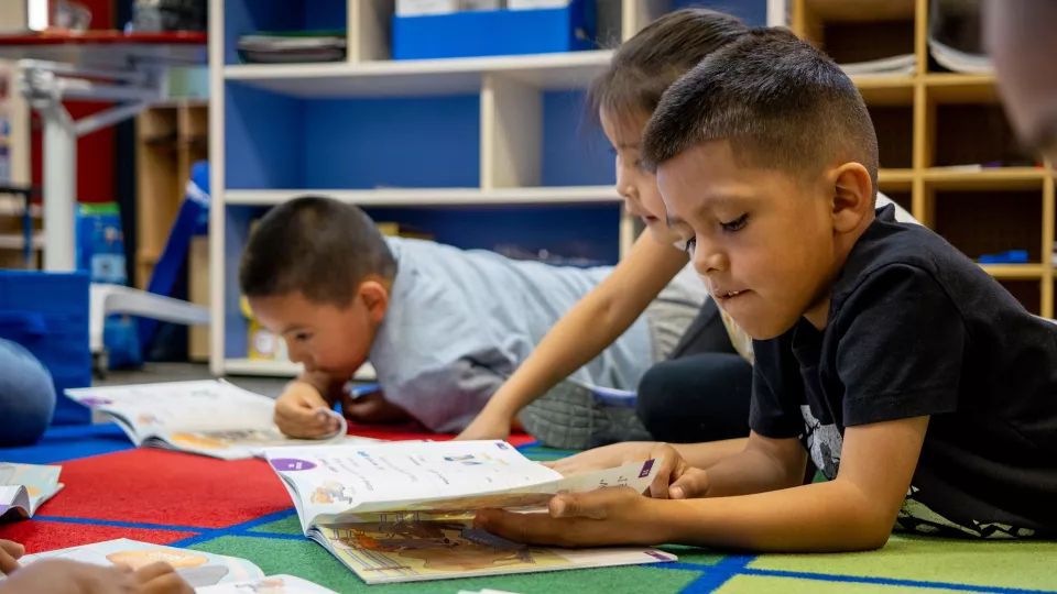 A young student enjoys reading a book while laying on a rug in a classroom.