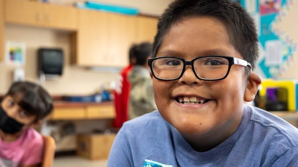 A student wearing black glasses sits in a classroom and smiles at the camera.