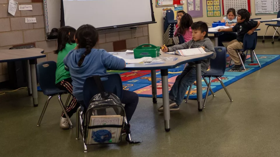 Students sit in groups at tables and work in their workbooks in a brightly colored classroom.