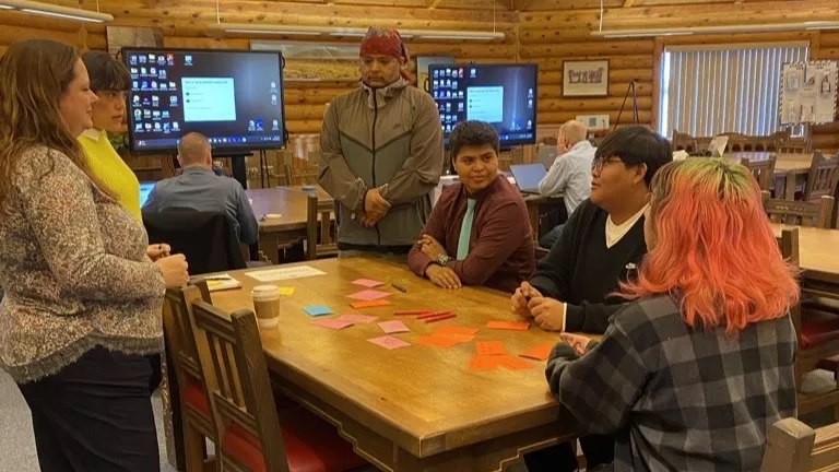 A group of students sit and stand around a table with stacks of index cards in various warm colors and talk to each other.