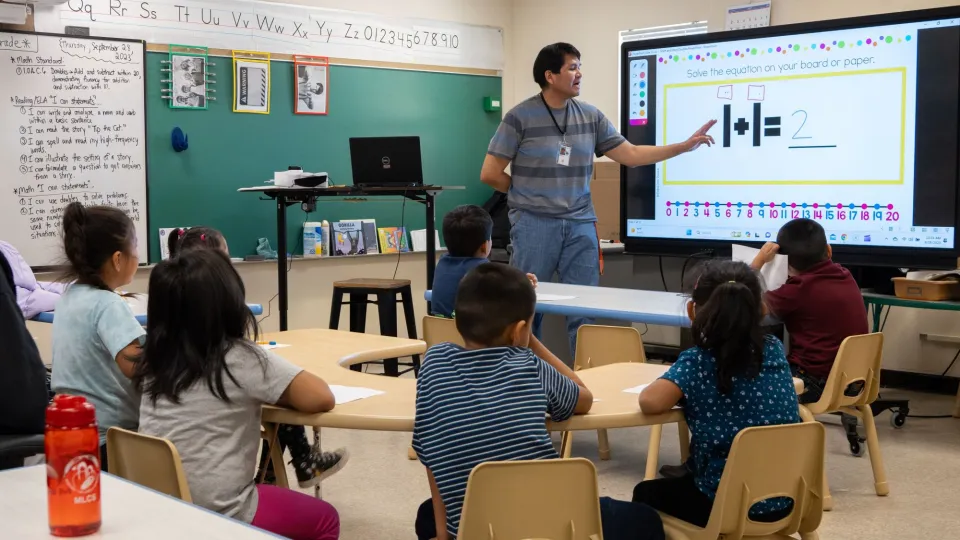 A teacher stands in front of a classroom of young students and teaches a math lesson using a smartscreen.