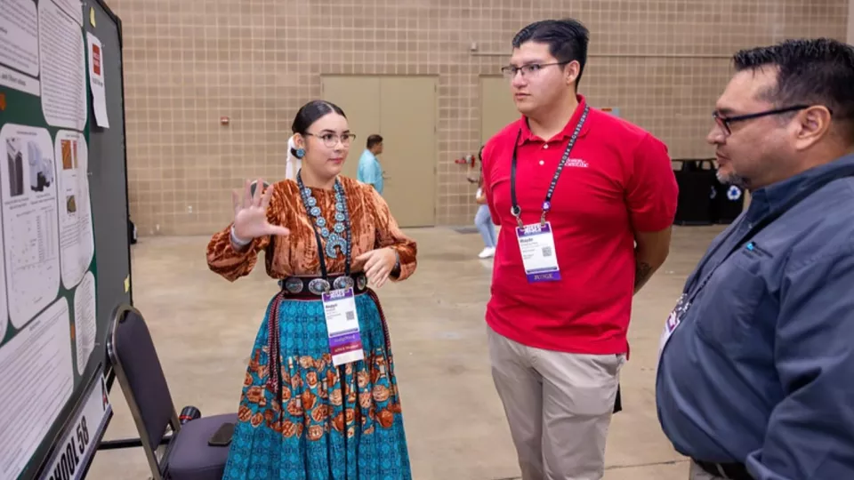 A student in a ribbon skirt and turquoise jewelry shows her research laid out on a cork board to two other people.
