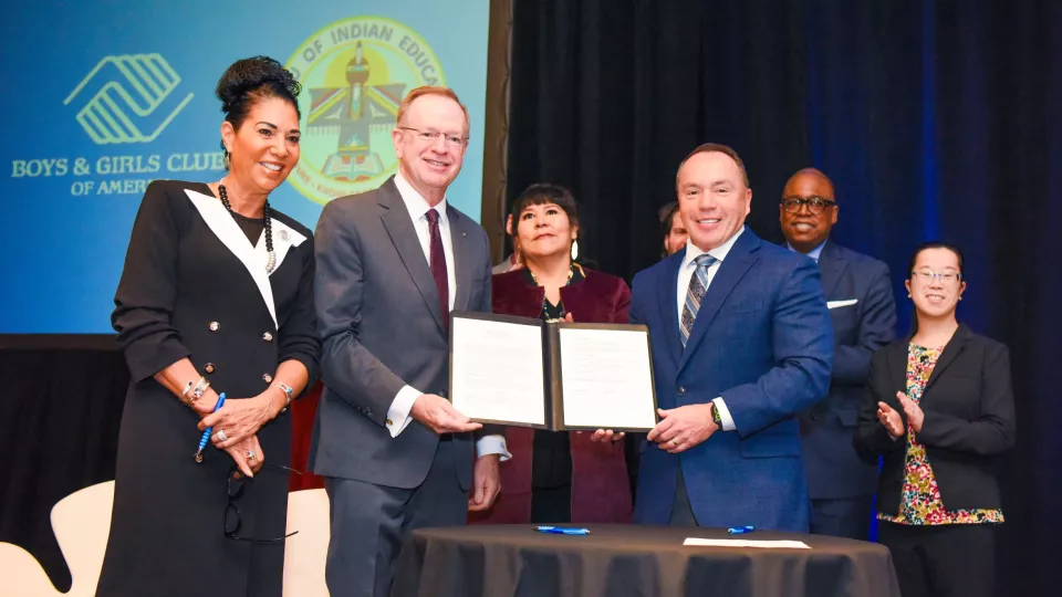 A group of people in business attire stand on stage and hold up a signed agreement.