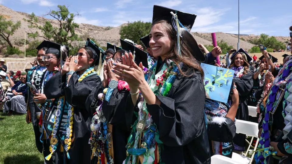 Students in black graduation robes and caps stand and clap in a celebratory outdoor setting.