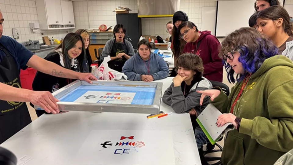 A group of art students surround a teacher at a table showing a demonstration.