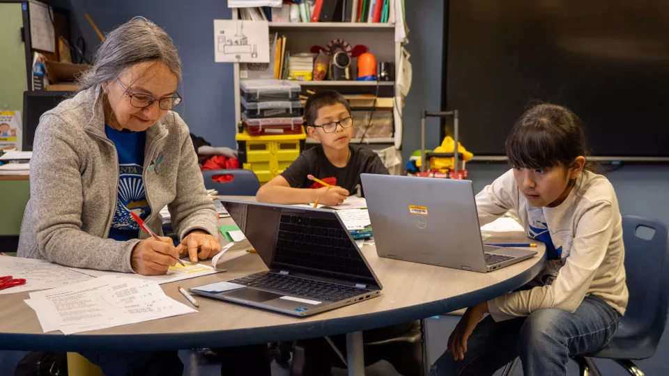 A teacher sits with two students at a table as they work on their laptops.