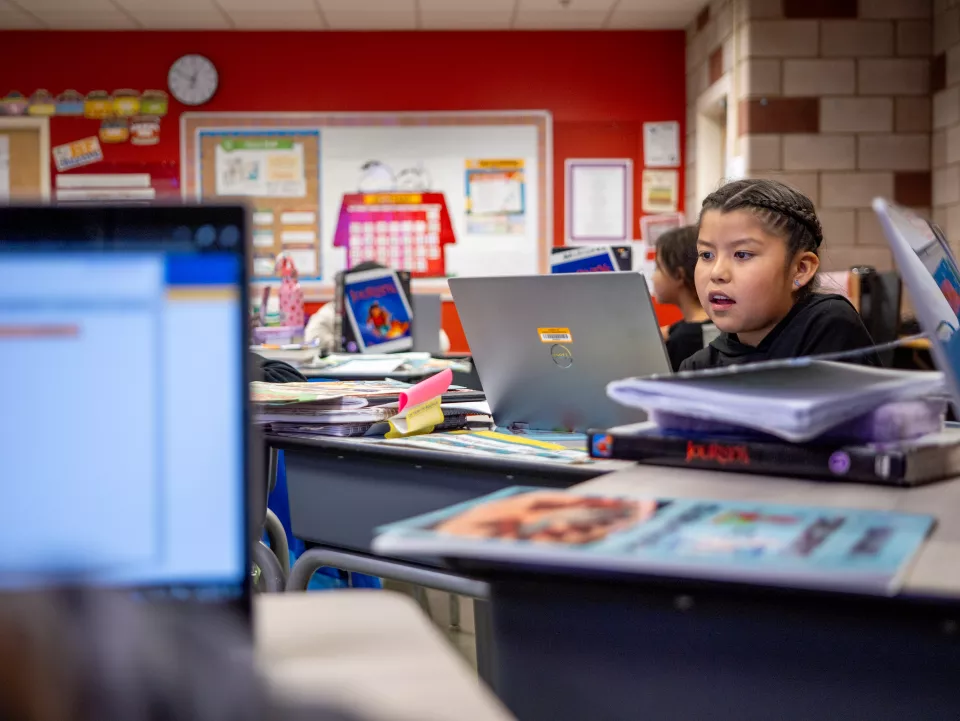 A young Indigenous student with braided hair sits at a desk in a classroom, working on a laptop. She appears focused and engaged. The classroom is filled with books, binders, and school supplies on desks. Other students are visible in the background, also working on laptops. The walls are decorated with educational materials, including a calendar and posters.