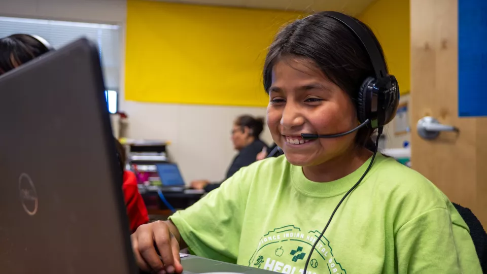 A student wearing head phones smiles while looking at her computer screen.