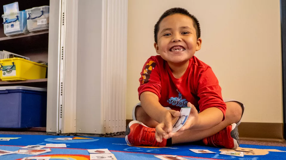 A young student in a red shirt and shoes sits on a brightly colored rug in a classroom and smiles.