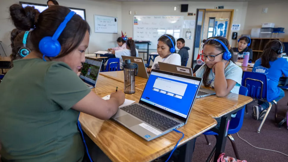 Students sit at their desks with headphones on working on laptops.