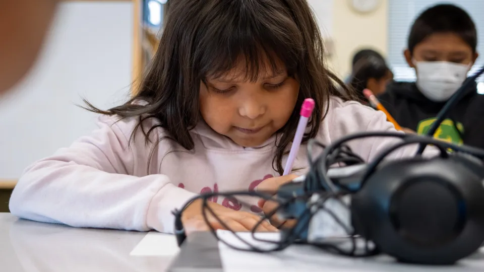 A student sits at a desk and writes on a piece of paper with a pencil with headphones in front of her.